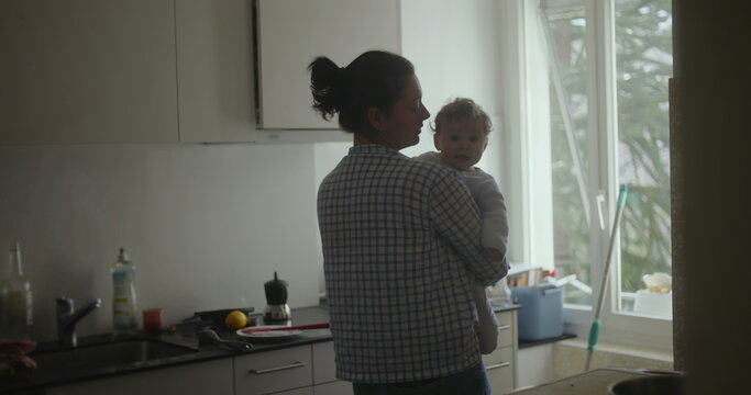 Mother holding baby in kitchen near window, morning domestic life scene showing tenderness, joy, and emotional bond between parent and child in everyday family moments
