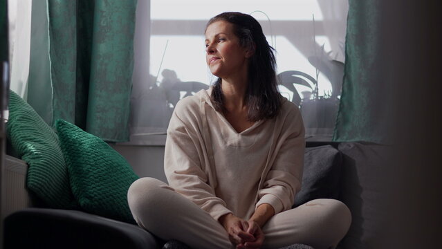 Middle aged woman sitting cross legged on sofa by window in soft daylight appearing thoughtful and distant in quiet introspective moment of solitude and reflection