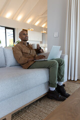 Obraz premium African American man wearing brown shirt holding white mug, typing on laptop at home on sofa