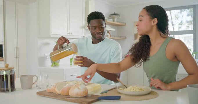African American couple making breakfast at kitchen island, he pouring OJ while she buttering bread
