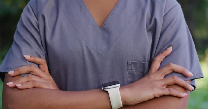 African woman arms crossed in yard, camera pulling back showing scrub top, watch, smiling greeting