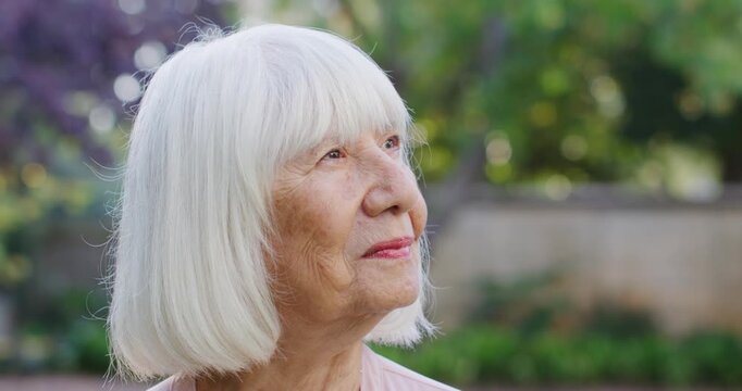 Senior Asian woman gazing upward, responding to sunlight and smiling in garden with beaded necklace