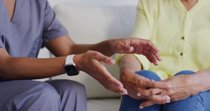 African American caregiver in scrubs rubbing then massaging senior woman's hands on sofa for rehab