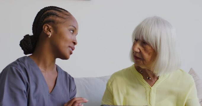 African American caregiver in scrubs leaning and explaining, soothing senior on couch with tablet