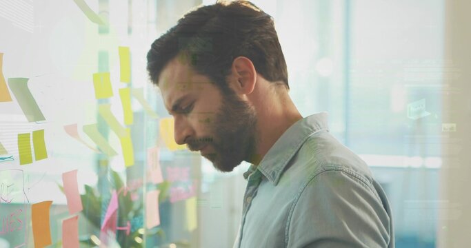 Studying man wearing light blue chambray shirt facing glass wall at office, with sticky notes