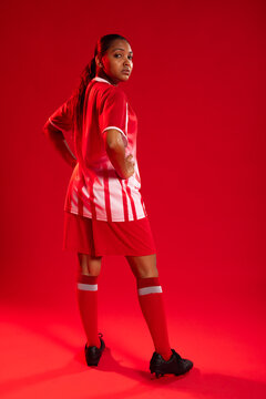 Woman posing in studio with red backdrop wearing soccer jersey, red shorts, cleats, socks, ponytail