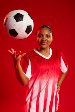 African American female in red white jersey posing in studio, tossing patterned soccer ball