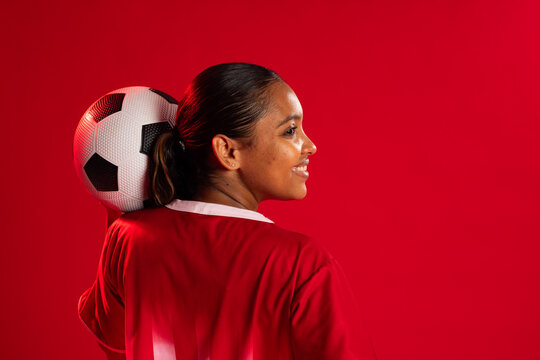 African American woman posing in red soccer jersey, cradling soccer ball on shoulder in studio