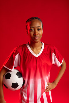 Woman posing with black-and-white soccer ball, wearing red-and-white jersey in studio red backdrop