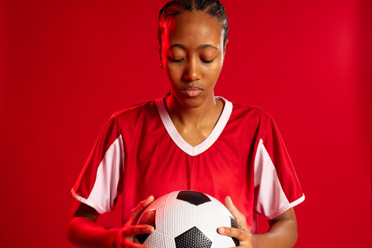 Adult African woman holding black-and-white soccer ball, wearing red and white jersey in studio