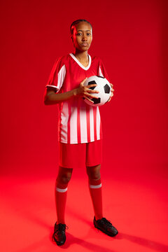 Adult African woman player posing at center on red studio floor holding soccer-ball wearing kit
