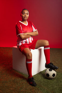 African woman sitting on white block in studio, wearing red jersey, resting on soccer ball