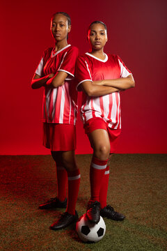 Female teammates posing on artificial turf in studio with soccer ball red jerseys red shorts cleats