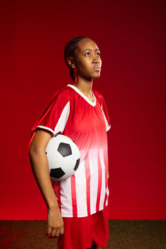 Adult African woman footballer posing holding soccer ball at hip on studio turf wearing red jersey