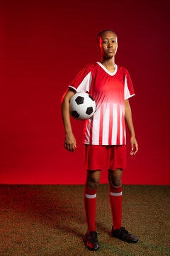 African American female soccer player in soccer kit standing on turf, red backdrop, holding ball