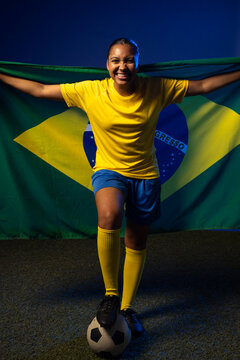 Female soccer player standing on turf wearing yellow jersey holding Brazilian flag and soccer ball