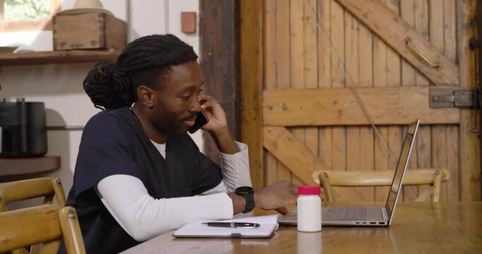 Taking call African American man in scrubs at table tapping laptop checking notes confirming med