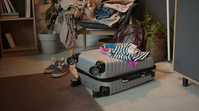 Medium no people shot of gray suitcase with wheels lying on floor in bedroom, travel carry-on backpack, pile of clothes on armchair, sneakers, at home of man or woman preparing to go on vacation