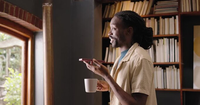 African man standing by arched window in shirt with smartphone, taking call and sipping white mug