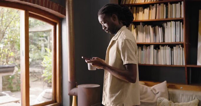Adult African man gazing out arched window holding white mug checking phone and speaking to someone