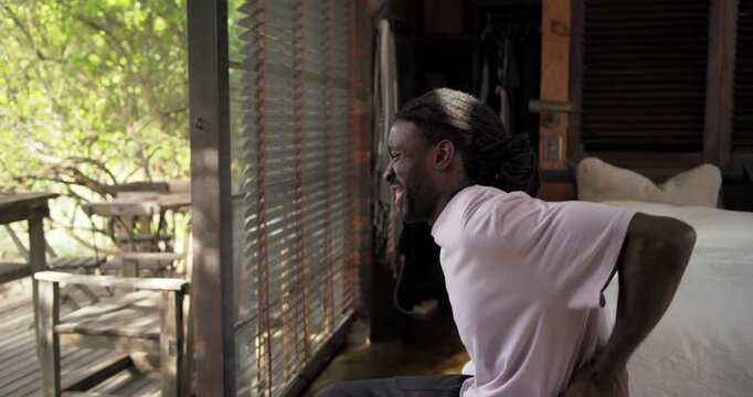 Dreadlocked African American man in pink tee rubbing lower back on bed relieving pain, copy space