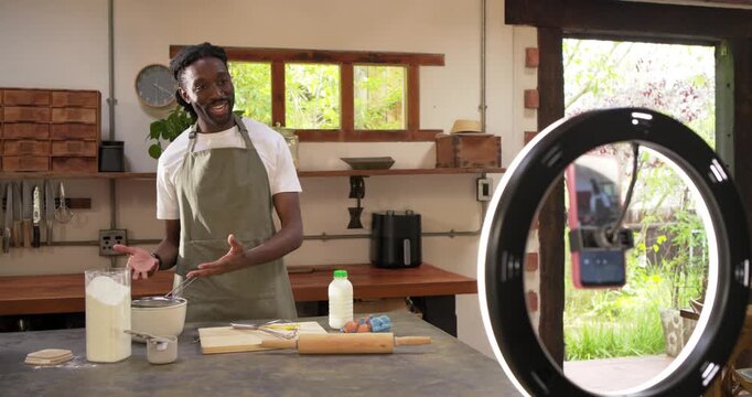 African American man in apron speaking to phone in ring light, recording recipe, measuring flour