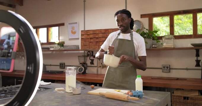 African man in green apron whisking bowl at kitchen island ring-light filming tutorial, copy space