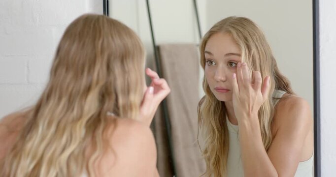 Woman checking reflection and smoothing under-eye skin with fingers at bathroom mirror for symmetry