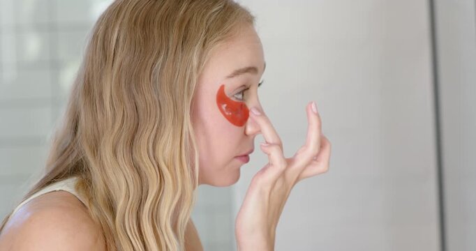 Female checking mirror and adjusting red under-eye gel patch at bathroom vanity, ensuring placement