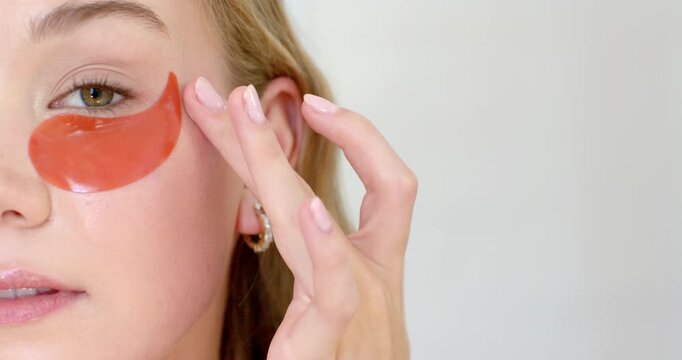 Adult female applying and smoothing red hydrogel patch under left eye using right hand, copy space