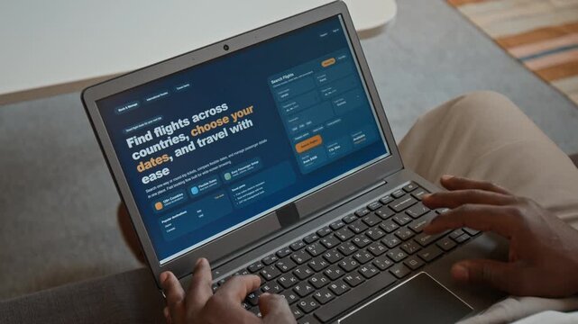 High-angle close-up of hands of anonymous black man sitting on couch at home, searching for flight tickets for family vacation on travel website, putting in dates and choosing destination in menu