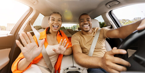 Young African American couple waving hands toward camera inside car, expressing excitement, greeting and joyful travel mood during a road trip experience © Anastasiya