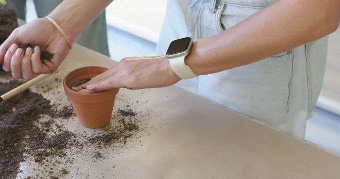 Female scooping, tamping soil from left pile at table, preparing terracotta pot for planting