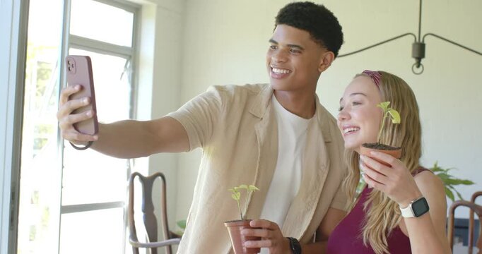 Diverse couple posing selfie in dining area, man extending smartphone, documenting seedling growth