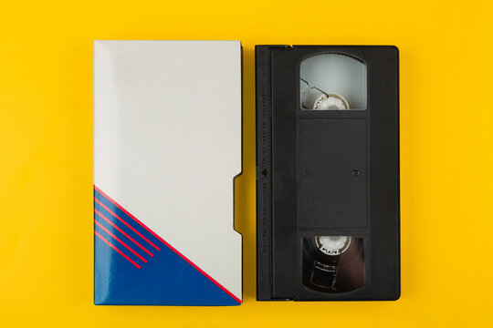A retro black VHS tape next to its cardboard sleeve on a bright yellow back