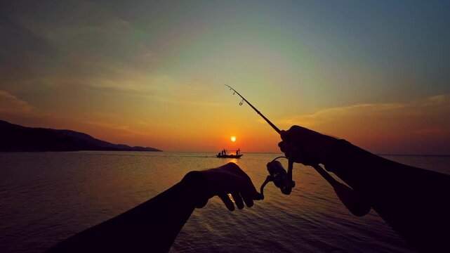 First person view of spinning rod directed toward horizon with small boat silhouette at sunset over sea, tranquil fishing scene and travel mood