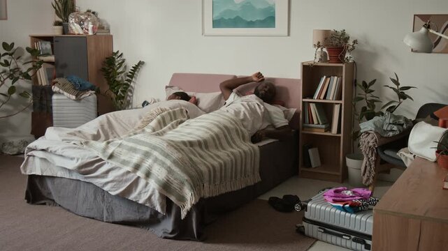 Full shot of young African American man waking up to alarm clock in morning, jumping out of bed, shaking up wife, both frantically packing clothes into suitcases to catch flight to go on vacation