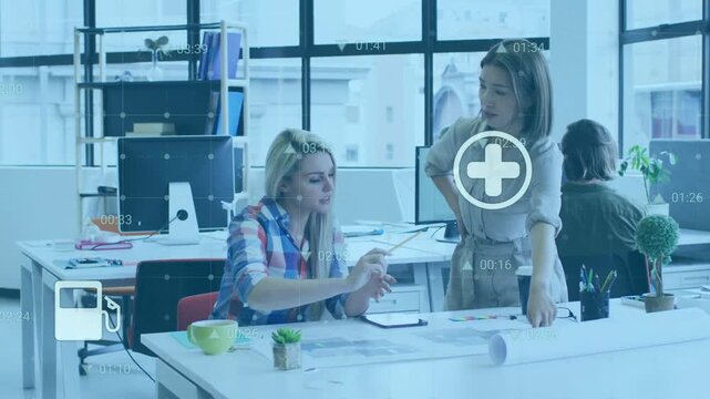 Two women collaborating at desk pointing and activating HUD icons while reviewing design layouts