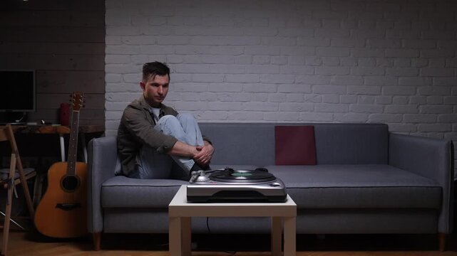 Man sitting on a sofa with his knees drawn up, looking thoughtful and melancholic while listening to music from a record player, surrounded by a guitar and a brick wall