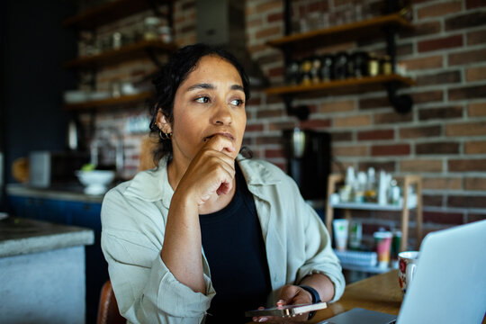 Thoughtful woman working from home in kitchen with smartphone and laptop