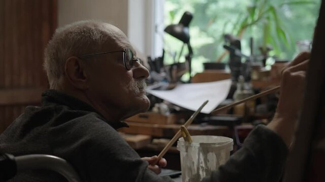 Senior artist on a wheelchair paints an oil painting on canvas in a studio