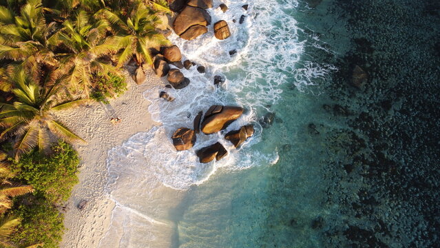 Top-down drone scene of waves washing over boulders and sandy coast with palm trees and clear turquoise water.
