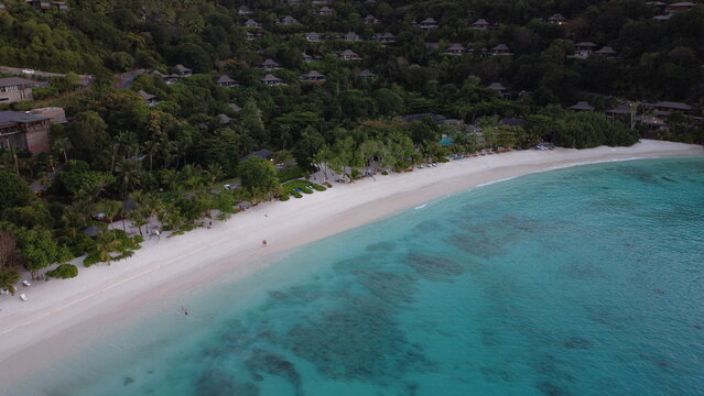 Drone shot of white sand shoreline with clear blue water and lush green forest, showing a secluded island coast from above.