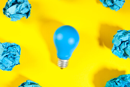 Blue light bulb surrounded by crumpled paper balls on yellow background