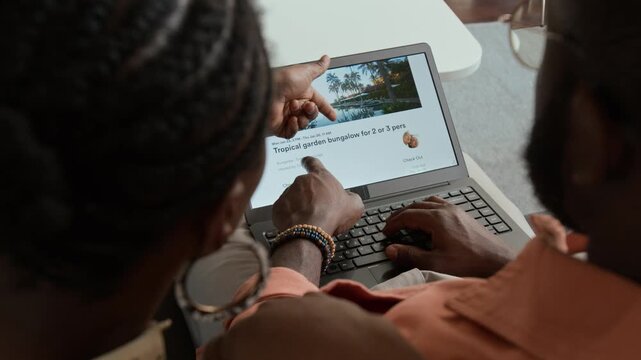 High-angle over-shoulder shot of young African American couple checking out exotic hotel for overseas vacation on travel booking agency website, pointing at amenities and discussing recreation options