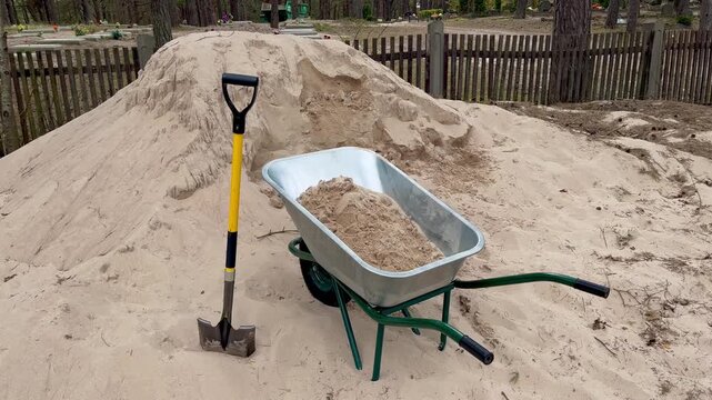 Pile of sand with shovel and wheelbarrow prepared near cemetery for leveling grave surface in spring showing maintenance work and seasonal care of burial site
