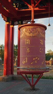Large red Buddhist prayer wheel with golden mantras under wooden pagoda. Warm sunset light creates spiritual, peaceful mood. Symbolizes meditation, religion, and Asian culture