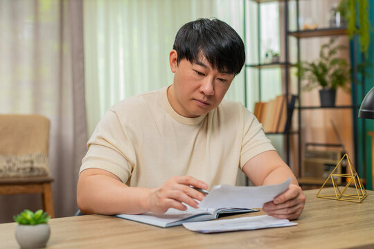 Asian man sitting at home table reviews utility bills, frowning, jotting notes in notebook, calculating amounts and deadlines. Chinese guy focuses tightly, tracking each figure for accurate budget.