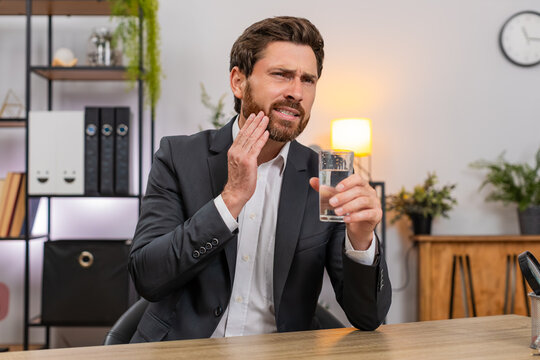 Middle-aged businessman at home office drinks cold water feels sharp tooth pain and searches dentist on smartphone. Freelancer guy at table presses cheek sighs hoping to book urgent clinic visit soon.