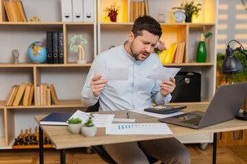 Fototapeta premium Young man on home table checks bills with tense lips and helpless hands after unexpected payment shock. Adult guy on home table tries to review costs and manage budget stress during home money crisis.